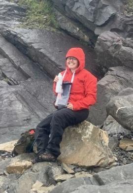 A woman posing for a photo outside, sitting on a rock formation, smiling, while holding a water bottle.