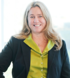 Susan wears a green shirt and black blazer. She has long blonde hair.